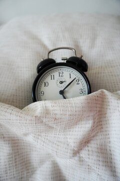 Black analog alarm clock under a blanket Close-up, soft light, fabric texture. Topic: morning, daily routine. The black vintage alarm clock is asleep. Vertical image.