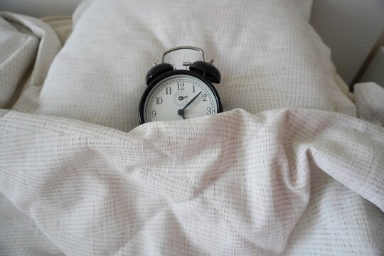 Black analog alarm clock under a blanket close-up, soft light, fabric texture. Topic: morning, daily routine. The black vintage alarm clock is asleep. Horizontal image.