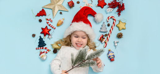 A happy 3-4-year-old girl in a hat lies on a blue background surrounded by Christmas toys and decor