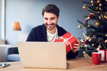 Happy young man opening Christmas gift during video call on laptop