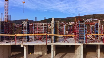 Workers are busy at a construction site where concrete walls are being erected. The sky is clear with a few clouds, and hills are visible in the distance beyond the structures.