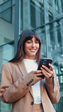 Young Caucasian businesswoman standing outside and smiling while using smartphone, with modern glass office building in the background