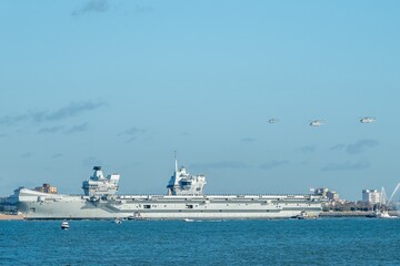 Portsmouth England -  November 30 2025: HMS The Prince of Wales returning to HMNB Portsmouth accompanies by the ports tug fleet and two Merlins and a Wildcat