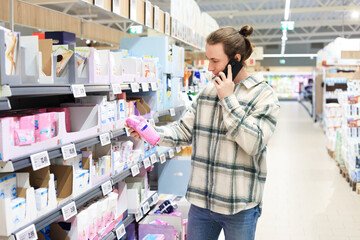 Obraz premium Young man talking on the phone while choosing feminine hygiene products in a supermarket aisle.