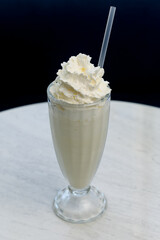 Vanilla milkshake with whipped cream in a tall glass on a white marble table.