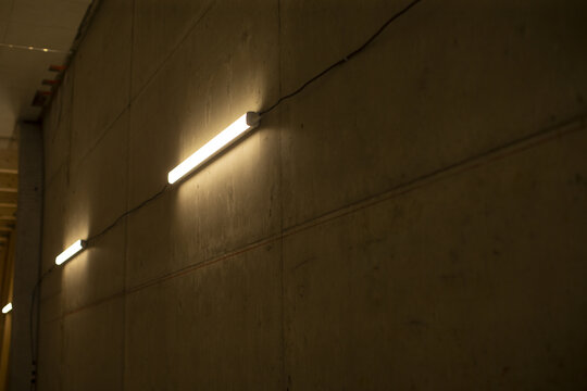 fluorescent tube lights on concrete wall in dim corridor, linear fixtures cast soft pools of light on raw texture, utilitarian