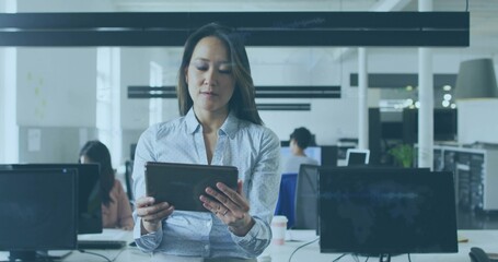 Holding tablet, woman in light blue patterned shirt standing at open-plan office desk with monitors