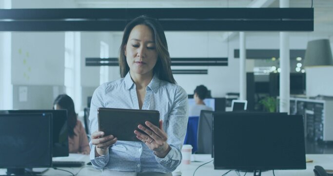 Standing central woman holding tablet, wearing light shirt in open-plan office with monitors