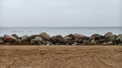 A structure made of large piled rocks protecting the beach on the shore of a large lake. Gray sky and gray water in inclement weather. Background. © Анатолий Еремин