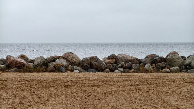 A structure made of large piled rocks protecting the beach on the shore of a large lake. Gray sky and gray water in inclement weather. Background.