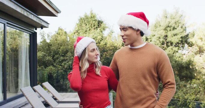 Receiving holiday cheer, diverse couple wearing Santa hats on deck brushing hair greeting camera