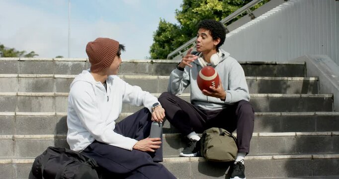 Diverse male friends grabbing football and explaining points while friend listening on bleachers
