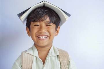 Close-up of a young boy smiling broadly while balancing a book on his head and wearing a backpack, isolated on white background. Education, learning, and school concept