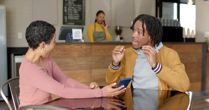 Diverse friends at cafe table, woman showing smartphone screen causing man laughing bumping fists