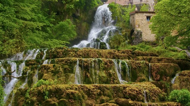 Vista amplia de la cascada principal de Orbaneja del Castillo, mostrando sus formaciones rocosas escalonadas cubiertas de musgo y el agua fluyendo entre la vegetaci&oacute;n verde del entorno.