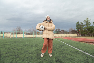 Portrait of a cheerful young female holding soccer ball at stadium in cloudy autumn weather