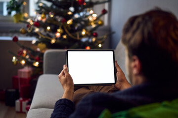Mock-up of young man holding digital tablet with blank empty screen during Christmas season while sitting on sofa