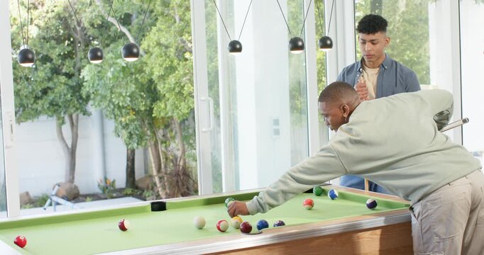 Diverse male friends striking cue ball at pool table scattering balls while preparing next shot