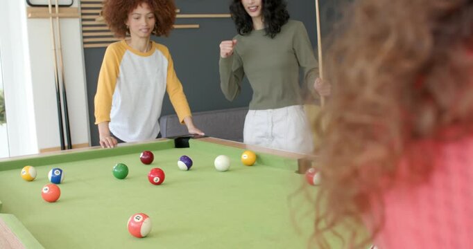After cue stick striking white ball, diverse female friends cheering in lounge by green-felt table