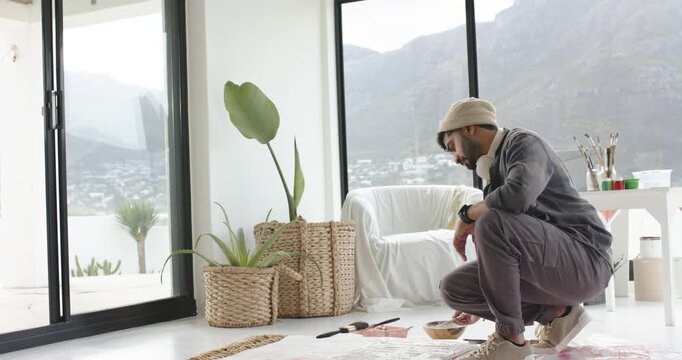 Indian man crouching at home grabbing paintbrush, dipping into paint, refining canvas, copy space