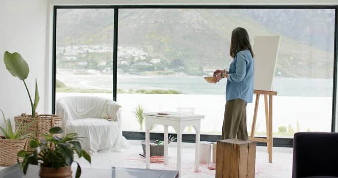Ocean view inspiring painter adjusting palette and inspecting canvas by window seeking inspiration