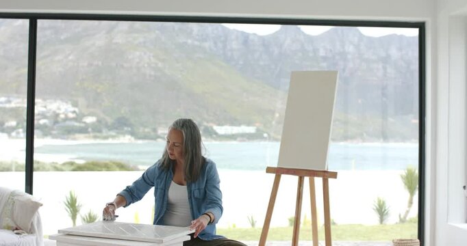 Mature White woman picking up primer jar and spatula, priming panel surface in coastal home
