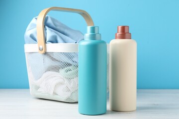 Laundry basket with clothes and bottles of detergent on white wooden table against light blue background, closeup
