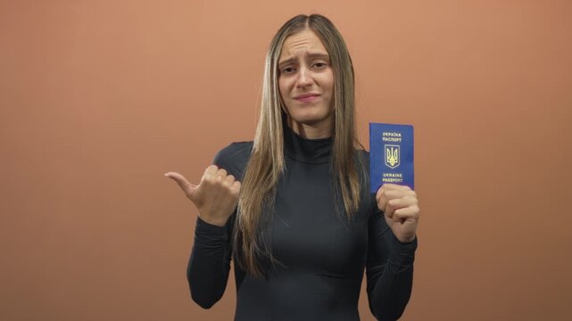 Young hispanic woman holding ukraine passport and pointing thumb to side in studio; identity travel skepticism.