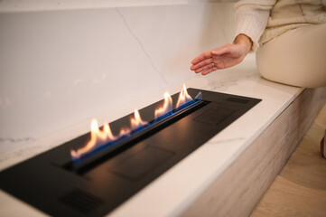 Obraz premium Woman warming her hands by the marble fireplace. Burning eco bio ethanol fireplace built into white marble. Automatic tabletop biofireplace. Minimalist interior, alternative bio fireplot fireplace.