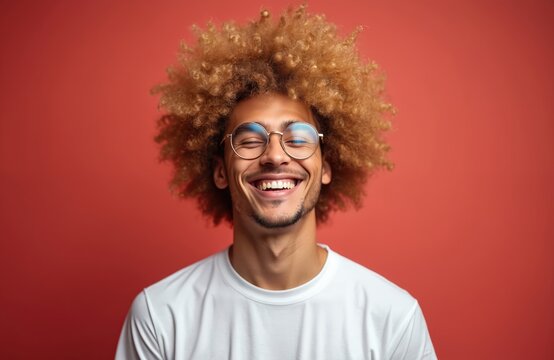 Happy mixed race guy with big blond afro hair smiles broadly. Handsome young man in glasses laughs showing white teeth. Cheerful student positive emotion, joy, fun on face isolated on red background - Powered by Adobe