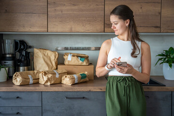 Young woman checks grocery delivery using a mobile app while standing in the kitchen. Concept of app-based meal kit subscription and digital grocery planning for home cooking.