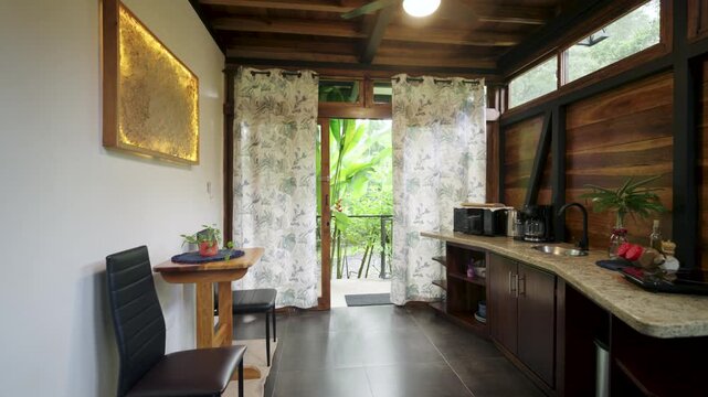 Interior of a rustic and modern cabin room featuring a wooden design, a kitchenette, and an open door revealing a lush green tropical jungle