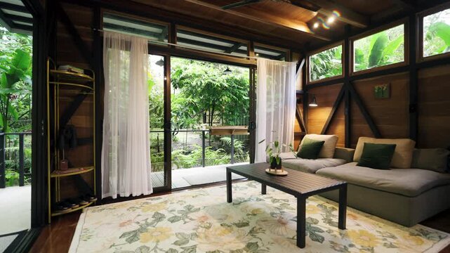 Comfortable living room interior of a modern cabin with wooden walls, large windows, and a view of the lush tropical garden outside
