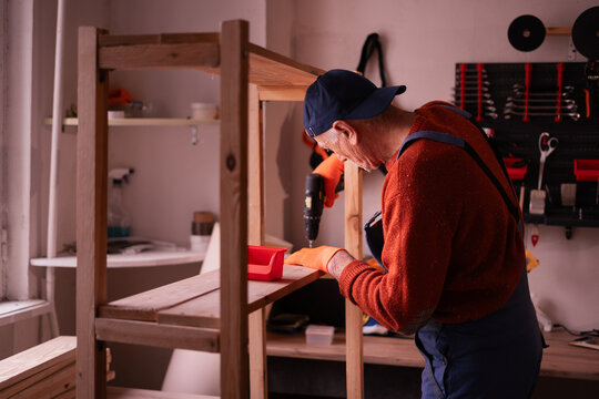 Senior Elderly Carpenter in blue overalls working in workshop. Craftsman assembling wooden shelf using electric drill drilling hole in carpenter's shop, making furniture from wood. - Powered by Adobe