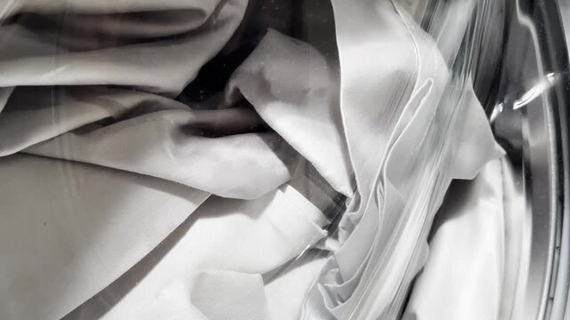 A closeup view of light white textiles inside the washing machine shows the fabric swirling behind