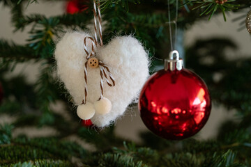 Soft felt heart with twine and pom‑poms hangs beside a glossy red bauble; selective focus and soft bokeh create a cozy holiday background for Christmas elements and happy new year cards.