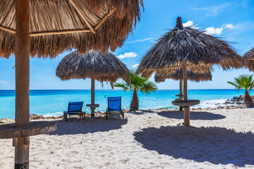 Beautiful view of Eagle Beach with palm leaf umbrellas and two loungers placed on sand near turquoise Caribbean sea. Aruba.