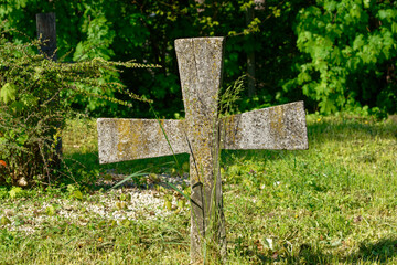 A weathered concrete cross stands in a peaceful grassy cemetery surrounded by nature and greenery.