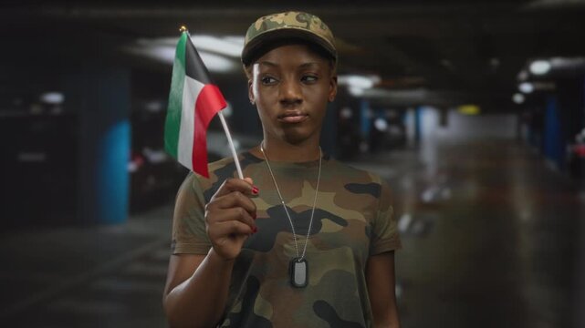 Young woman holds kuwait flag on a stick while showing dogtags in camouflage uniform and cap inside a building; solidarity support pride.