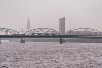 Train Crossing the Railway Bridge Over the Daugava River with TV Tower and Modern Skyline in Winter Fog – 08.12.2025. Riga. Latvia