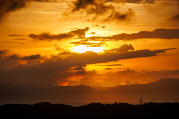 Dramatic sky with layered clouds illuminated by warm orange and yellow hues as the sun sets behind the distant hills and ocean