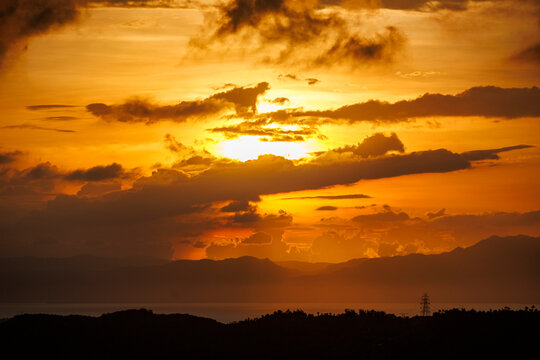 Dramatic sky with layered clouds illuminated by warm orange and yellow hues as the sun sets behind the distant hills and ocean
