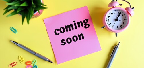 Pink sticky note says coming soon on yellow desk surface. Alarm clock, pen, paper clips, and green plant are nearby. Prepare for new launch or event.