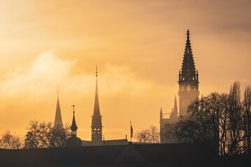 The evening sun casts an orange glow on the clouds, turning the roof of the Steigenberger Hotel and the Cathedral "M&uuml;nster unserer lieben Frau" into silhouettes. Konstanz, Baden-W&uuml;rttemberg, Germany.