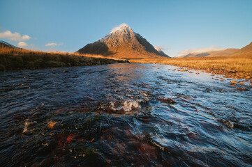 Scenic morning view of Buachaille Etive Mor in Glencoe, Scotland, with a flowing river in the foreground and clear blue sky. Natural landscape, no people