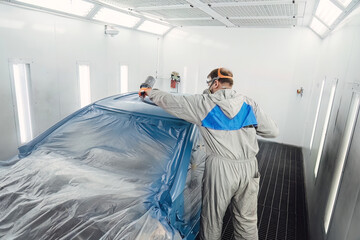 Professional painter in a protective suit spraying paint onto the car body inside a clean, modern spray booth. High quality automotive repair and bodywork service maintenance for vehicles.
