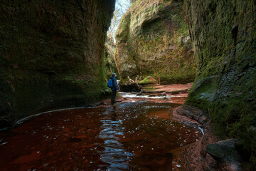 Hiker standing in the red-water stream of Finnich Glen, Scotland, surrounded by moss-covered sandstone walls and dramatic canyon scenery. Outdoor adventure landscape.