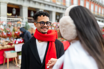 Happy young man wearing red scarf and glasses interacting with woman at christmas market in plaza mayor