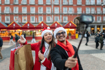 Happy couple wearing santa hats making a selfie with a action camera at christmas market in plaza mayor, madrid
