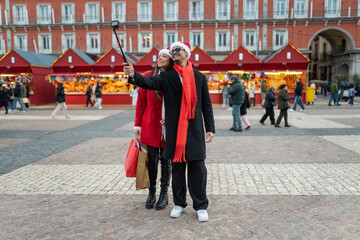 Happy couple wearing santa hats taking selfie with camera on a stick, walking through christmas market